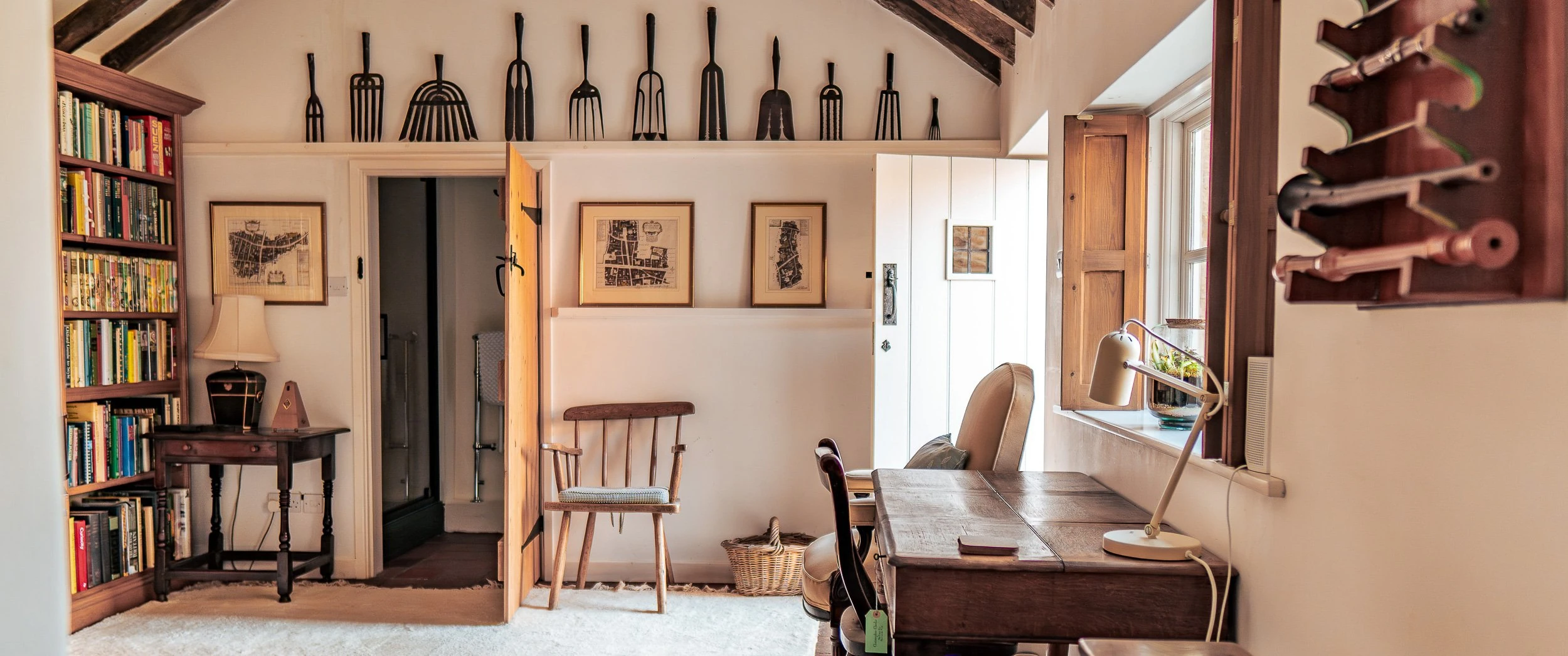 Book room at Well Cottage with fitted timber shelving, framed maps, a Windsor-style armchair and antique hay forks ranged along the wall beneath the pitched ceiling.