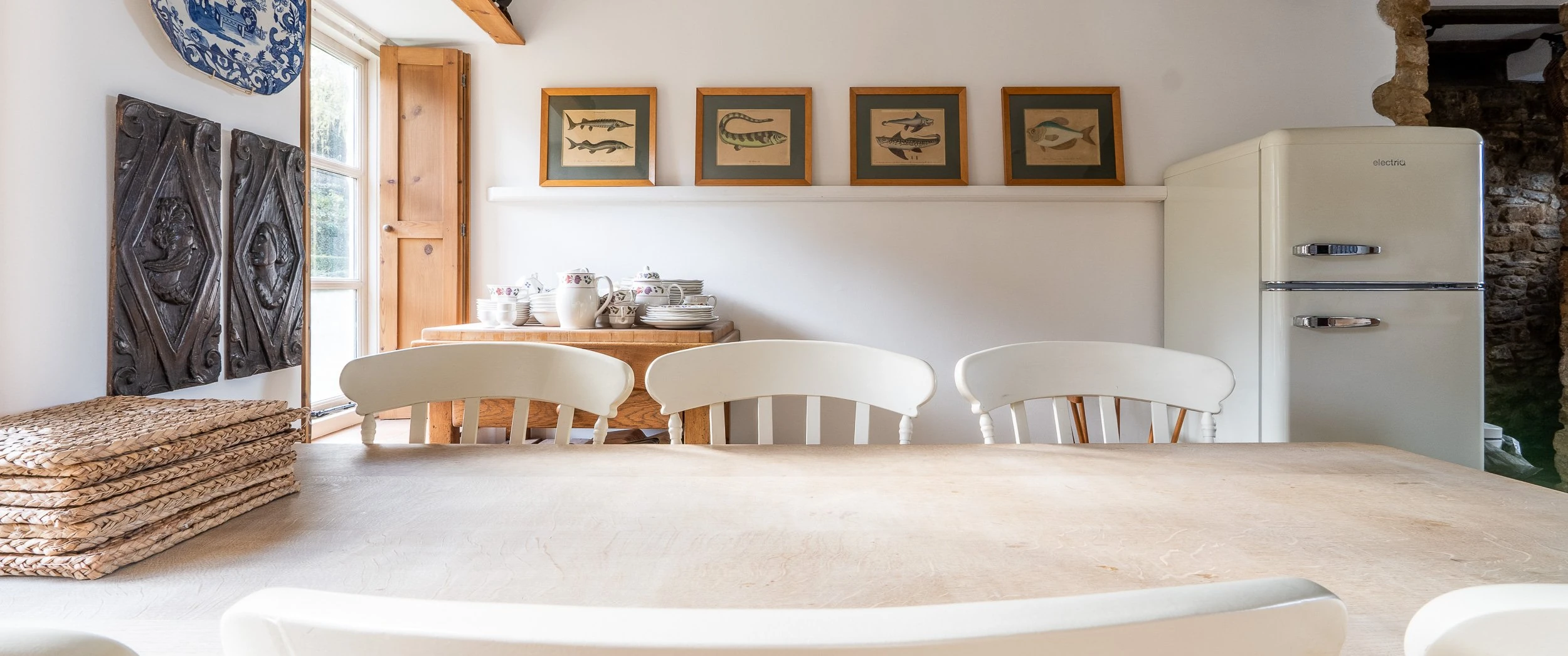 Kitchen at Well Cottage seen across the pale timber table, with painted spindle-back chairs, framed fish prints on a shelf, carved oak panels and a cream fridge against the exposed stone.