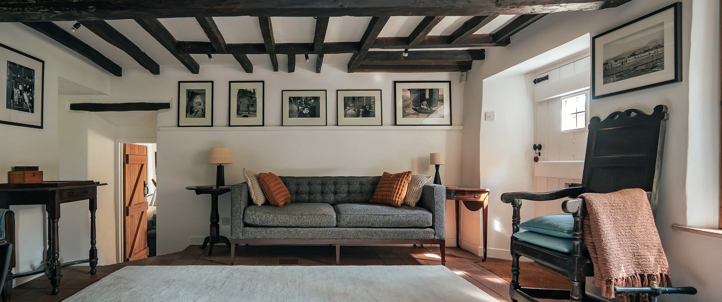 Beamed reception room at Well Cottage, Ledwell, with a buttoned grey sofa, framed black-and-white photographs on a picture shelf and an antique wainscot armchair on tiled floor.