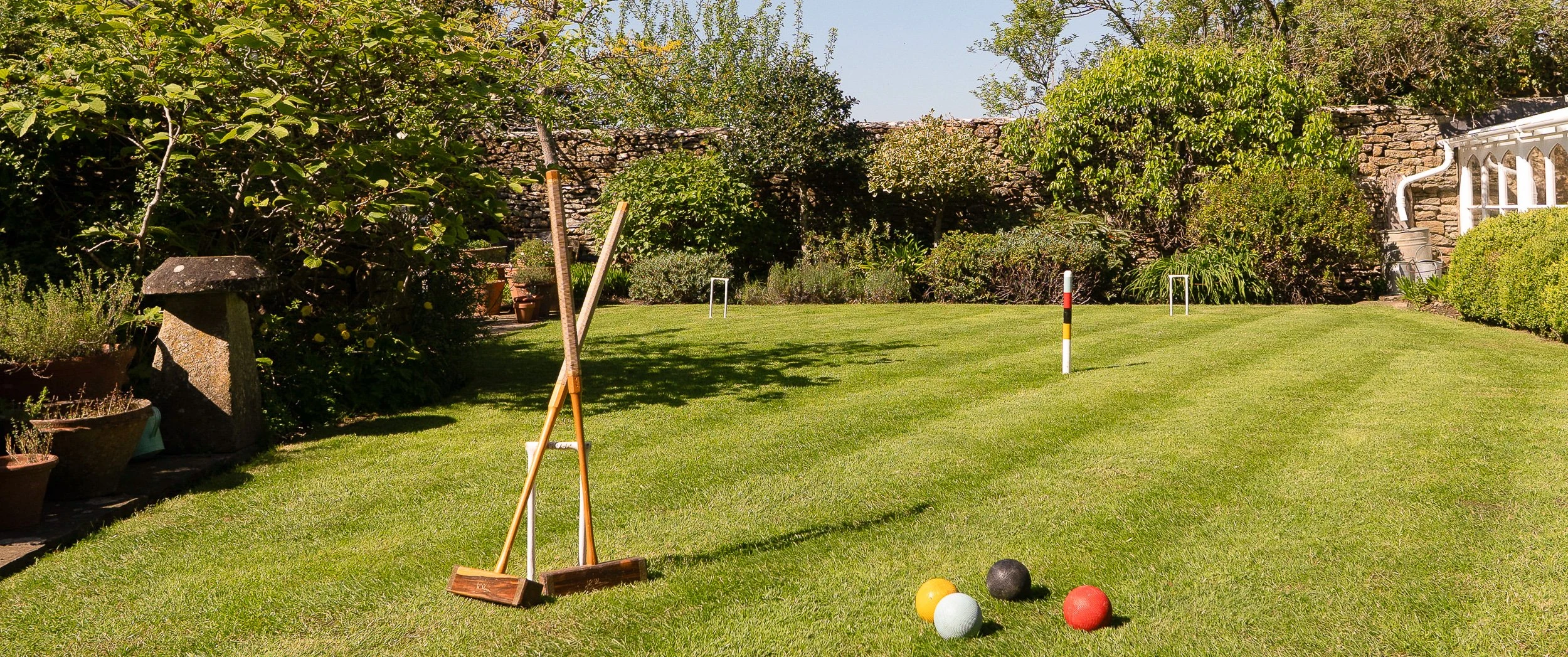 Level croquet lawn at Well Cottage set with hoops, a striped peg, mallets and coloured balls, enclosed by a dry stone wall, planted beds and a glasshouse.