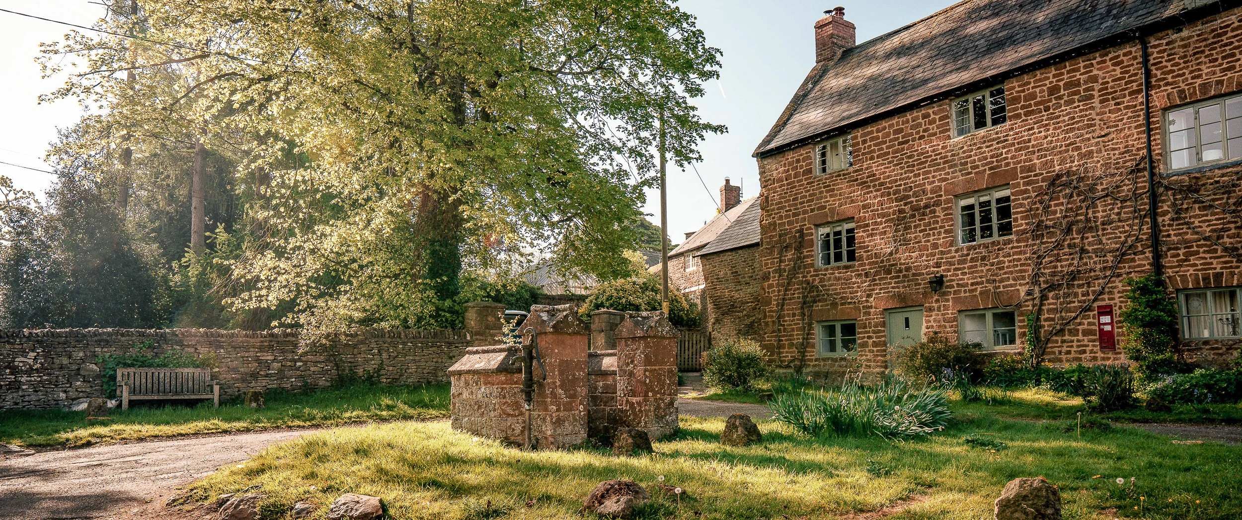 Stone well head with an iron pump on the green at Ledwell, with an ironstone cottage range behind and a wooden bench against the boundary wall.