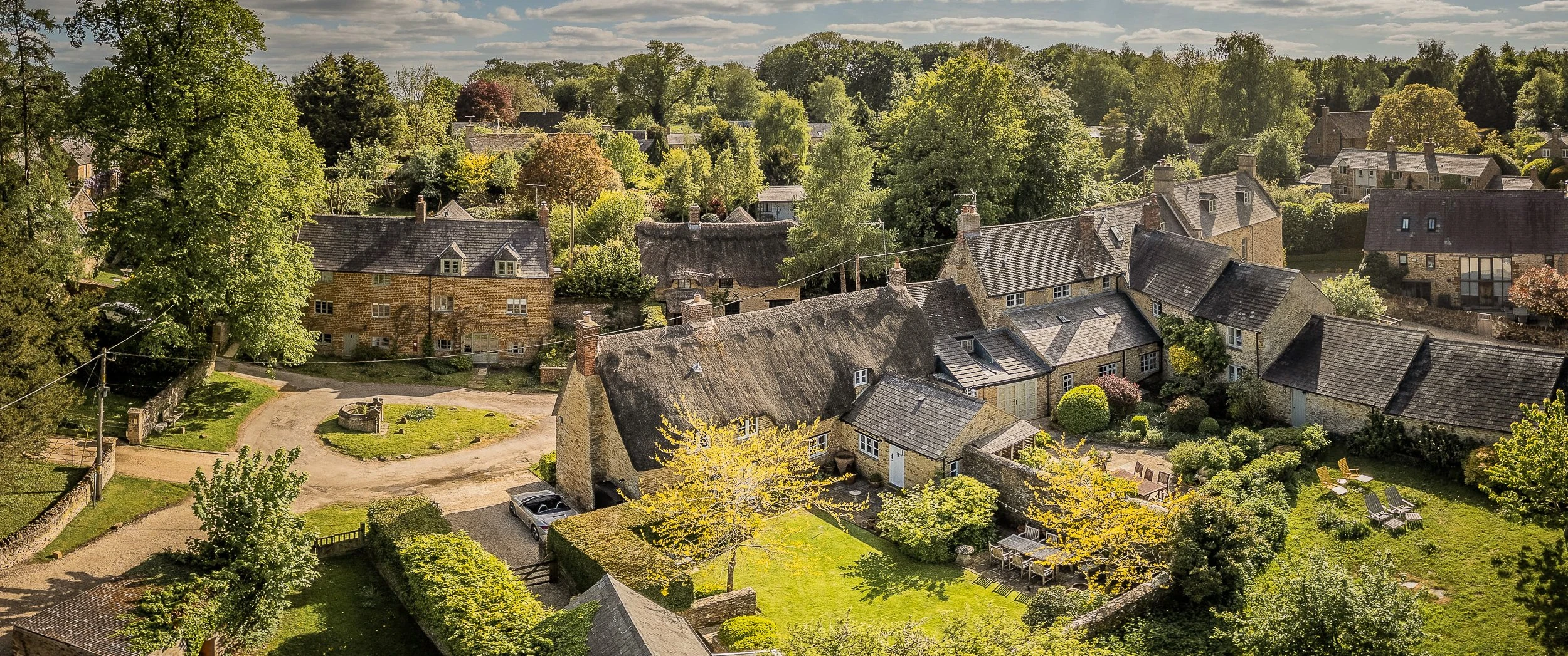 Aerial view over the thatched roof, walled garden and stone outbuildings of Well Cottage, Ledwell, with the village lane and well head beyond.
