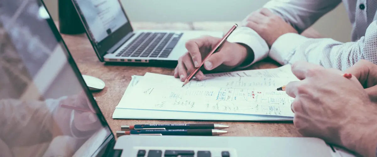 Hands with pencil over notes beside laptops, project management of bespoke hospitality furniture